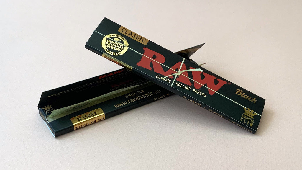 RAW Black - Classic King Size Slim, featuring ultra-thin, unbleached, and double-pressed artisan paper with a tan black and gold red themed packaging. RAW King Size Slim natural unrefined, 110x44mm, 32 leaves per booklet.