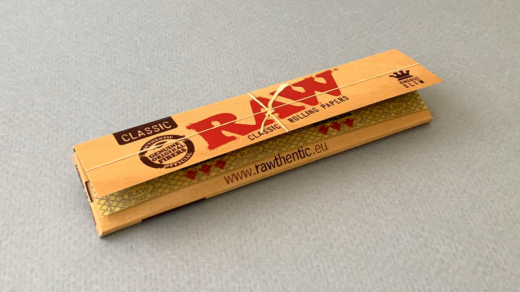 RAW Black - Classic King Size Slim, featuring ultra-thin, unbleached, and double-pressed artisan paper with a tan black and gold red themed packaging. RAW King Size Slim natural unrefined, 110x44mm, 32 leaves per booklet.